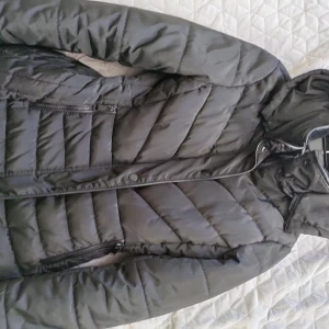 Svart pufferjacka med huva - Säljer en svart pufferjacka med huva och mjukt foder. Jackan har dragkedja, knappar och två fickor framtill. Snyggt quiltad design och elastisk midja för bättre passform. Perfekt för kalla dagar när du vill hålla dig varm och stilren. 