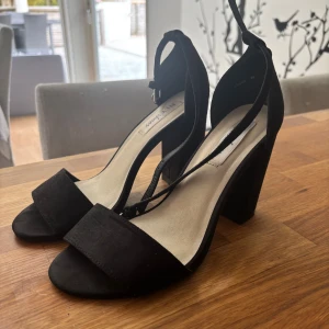 Svarta klackade sandaler från NLY Shoes - Stilrena svarta sandaler från NLY Shoes med bred rem över tårna, öppen tå och chunky klack. Skorna har en smal rem runt vristen med spänne och är gjorda i mockaliknande material. Perfekta för att lyfta din outfit med en elegant touch.