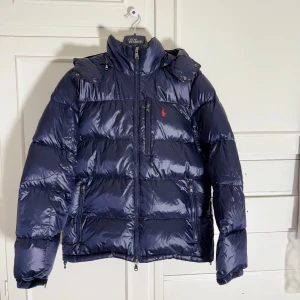 Mörkblå pufferjacka från Polo Ralph Lauren - Säljer en mörkblå pufferjacka från Polo Ralph Lauren med glansig finish och röd logga på bröstet. Jackan har huva, tre dragkedjefickor framtill och kraftig vaddering som håller dig varm. Perfekt för kalla dagar och riktigt snygg till jeans.