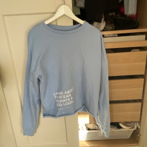 Ljusblå sweatshirt från Organ Cartel - Ljusblå sweatshirt med vit text framtill: 'Caring about the ocean is always a good look.' Tröjan har rund hals, långa ärmar och ribbade muddar. Perfekt för en chill och avslappnad stil.