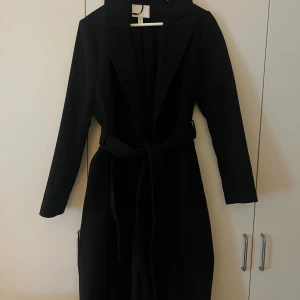 Svart kappa med bälte från H&M - Stilren svart kappa från H&M med klassisk krage och bälte i midjan. Lång modell med raka ärmar och snygg passform. Perfekt för dig som vill ha en tidlös look under höst och vinter. Materialet är mjukt och känns lyxigt.