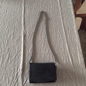 Svart axelväska med kedjerem - Stilren svart axelväska i skinn med silverfärgad kedjerem och magnetlås. Insidan är fodrad i rött vilket ger en snygg kontrast. Perfekt storlek för det viktigaste och passar till många olika outfits. 