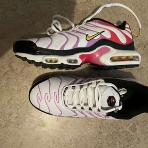 Nike Air Max Plus vita/lila/röda - Nike Air Max Plus sneakers i vitt med lila vågiga detaljer och röda inslag på sidorna. Swoosh-logga i gult och svart, svart yttersula och synlig Air Max-dämpning. Ovandel i mesh och syntet, snörning framtill och svart innersula med gul Nike Air-tryck.