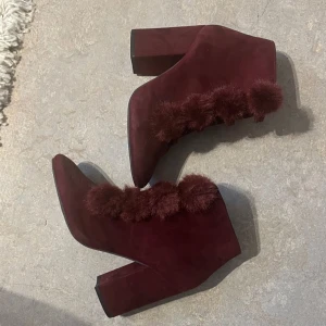 Vinröda mocka stövletter med pälsdetaljer - Säljer ett par vinröda stövletter i mocka med chunky klack och spetsig tå. Skorna har snörning framtill och är dekorerade med fluffiga pälsbollar längs snörningen för en unik och trendig look. Perfekta för dig som vill sticka ut med dina skor.