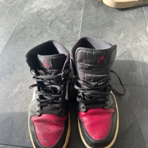 Nike Air Jordan 1 Mid Bred - Säljer ett par Nike Air Jordan 1 sneakers i svart, rött och vitt. Klassisk mid-top modell med snörning, perforerad tå och ikonisk Jumpman-logga på plösen. Snygg kontrast mellan färgerna och den röda yttersulan ger en riktigt fet look. Perfekta för dig som gillar streetwear.