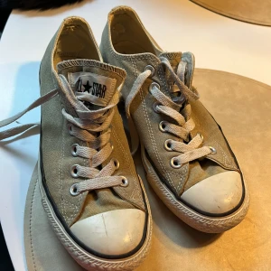 Beige Converse All Star sneakers - Säljer ett par klassiska Converse All Star sneakers i beige canvas med vita gummitår och sula. Skorna har snörning och låg modell, med den ikoniska All Star-loggan på plösen och hälen. Perfekta för en avslappnad och cool stil.