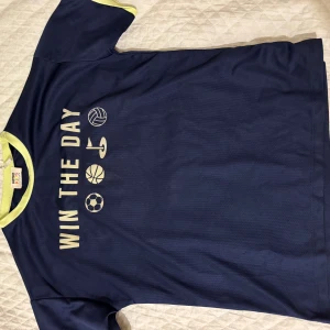 Blå sportig t-shirt WIN THE DAY - Sportig marinblå t-shirt från Koton Kids med trycket 'WIN THE DAY' och sportikoner på bröstet. T-shirten har korta ärmar och ljusgula detaljer vid halsringning och ärmslut. Perfekt för dig som gillar en aktiv stil.