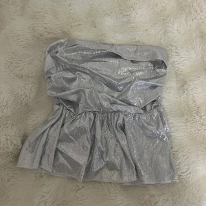 Glittrig silver tubtopp med volang - Säljer en glittrig silverfärgad tubtopp med volangkant nertill. Toppen har bandeau-modell och är axelbandslös med smockad baksida för extra komfort. Perfekt för dig som vill sticka ut med en metallic look.