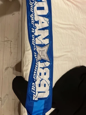 Blå fotbollshalsduk Djurgården LgS - Säljer en blå väst med breda ränder och vit text, inspirerad av Djurgårdens IF. Västen har ett sportigt utseende med tryckta bokstäver och DIF-emblem framtill. Materialet är troligtvis polyester och designen är loose fit, perfekt för att visa support på matchen.