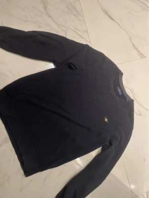 Mörkblå stickad tröja från GANT - Säljer en mörkblå stickad tröja från GANT i storlek M. Tröjan har rund hals med randig detalj, broderad GANT-logga på bröstet och långa ärmar. Perfekt till höst och vinter för en clean och klassisk look.