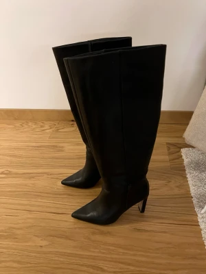 Svarta höga boots i skinn från NA-KD - Stilrena svarta boots i äkta skinn från NA-KD med högt skaft och spetsig tå. Skorna har en stadig klack och dragkedja på insidan för enkel påtagning. Perfekta för att lyfta din outfit med en edgy vibe. Klacken är 8 cm