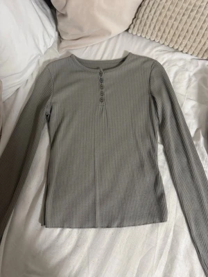 Grå ribbad långärmad topp - Säljer en grå ribbad långärmad topp med rund halsringning och knappar framtill. Toppen har en slim passform och är tillverkad i mjukt bomullsmaterial, perfekt för lager på lager eller att bära som den är.