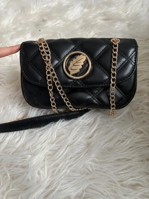 Svart  axelväska från ioaku  - Snygg svart axelväska från ioaku i quiltad skinnimitation med guldfärgade kedjedetaljer och ett stort lövformat metallogo på locket. Insidan är fodrad i rosa och har en praktisk innerficka med dragkedja. Perfekt storlek för det viktigaste.