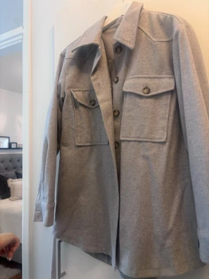 Grå overshirt med stora fickor - Snygg grå overshirt med stora bröstfickor och knappar framtill. Jackan har en rak passform, klassisk krage och långa ärmar. Perfekt att slänga över en hoodie eller t-shirt för en chill look.