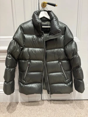 Mörkgrön dunjacka med dragkedja - Säljer en riktigt snygg mörkgrön pufferjacka med hög krage och två snedställda fickor med dragkedja. Jackan har en glansig finish och breda, quiltade partier som ger en cool och modern look. Perfekt för kalla dagar när du vill hålla stilen.