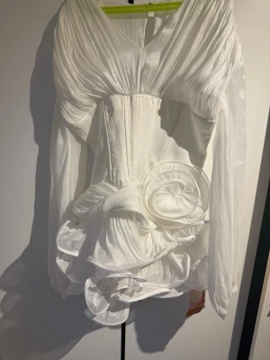 Vit kortklänning med volang och rosett - Supersnygg vit kortklänning med transparenta långärmar, markerad midja och stora volangdetaljer samt en dekorativ rosett framtill. Klänningen har en unik struktur och ger en riktigt statement-look. Perfekt för dig som vill sticka ut! Perfekt till studenten