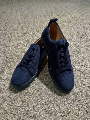 Marinblå Louboutins - Marinblå skor från Christian Louboutin i mjuk mocka med klassisk röd sula och broderad logga på hälen. Skorna har rund tå, snörning och platt sula. Insidan är beige och sulan har den ikoniska Louboutin-signaturen i rött.