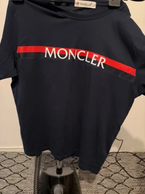 Mörkblå Moncler t-shirt med röd detalj - Snygg mörkblå t-shirt från Moncler med vit och röd logga över bröstet. Klassisk passform och rund hals. Tillverkad i mjuk bomull som känns skön mot huden. Perfekt för dig som vill ha en stilren och sportig look.
