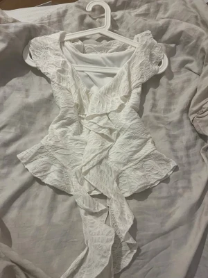 Vit omlottopp med volang från SHEIN - Supersöt vit omlottopp från SHEIN i storlek S. Toppen har volangdetaljer längs kanterna och ett lätt strukturerat tyg. Knytband i midjan och v-ringad framtill. Tillverkad i mjuk polyesterblandning med lite stretch för skön passform.