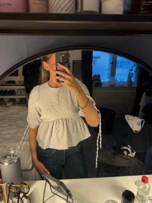 Vit blus med öppen rygg och knyten - Supersöt vit/grå blus i strukturerat tyg med puffiga halvlånga ärmar och volang nedtill. Blusen har öppen rygg med knytband både i nacken och i midjan samt knyt vid ärmsluten. Perfekt för dig som vill ha något unikt och trendigt.