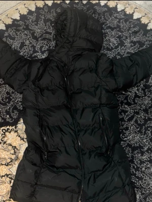 Svart lång pufferjacka med huva - Säljer en svart lång pufferjacka från USA Polo Assn. Jackan har en stor huva, dragkedja framtill och två sidofickor med dragkedja. Perfekt för kalla dagar och riktigt skön att bära. Klassisk look som funkar till det mesta.