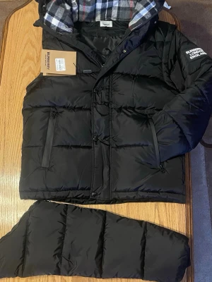 Svart pufferjacka från Burberry - Säljer en svart pufferjacka från Burberry med avtagbara ärmar och rutigt foder i huvan. Jackan har dragkedja, knappar och två fickor med dragkedja. Klassisk Burberry-logga på ärmen och riktigt schysst oversized look.