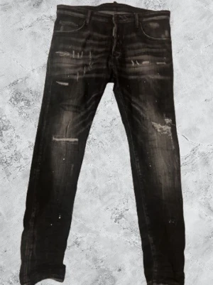 Svarta Dsquared2 jeans med slitningar - Säljer ett par svarta jeans från Dsquared2 med coola slitningar och distressed detaljer framtill. Jeansen har klassisk femficksdesign, raka ben och en patch bak på linningen. Materialet är jeans i bomull och passformen är normal.