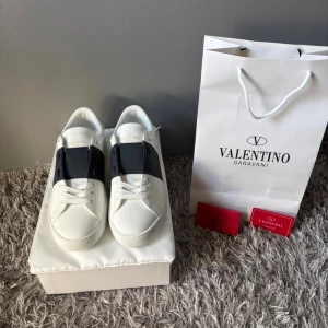 Valentino Garavani Open snears vit/blå - Snygga Valentino Garavani Open sneakers i vitt skinn med blå band över snörningen. Klassisk rund tå, platt sula och diskret guldlogga på plösen. Perfekta för dig som vill ha en clean och lyxig look med en modern twist.