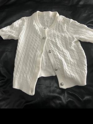 Vit kabelstickad kofta med knappar - Säljer en vit kabelstickad kofta med korta ärmar och silvriga knappar framtill. Tröjan har ribbade muddar vid ärmslut och nederkant samt en klassisk rund halsringning. Perfekt att slänga över en topp för en chill look.