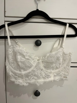 Vit spets bh från Lindex  - Säljer en vit spets bh från Lindex med justerbara axelband och hakknäppning bak. Bhn har vacker spets med blommigt mönster och en liten metallbricka med logga framtill. 