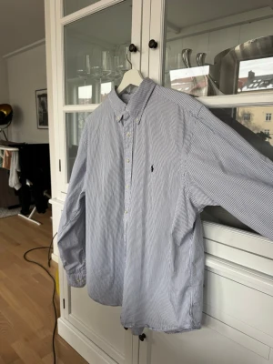 Randig skjorta från Ralph Lauren - Klassisk vit och blå randig skjorta från Ralph Lauren i modellen Classic Fit. Skjortan har button-down krage, långa ärmar och knappar framtill. Tillverkad i mjuk bomull och har den ikoniska loggan broderad på bröstet.