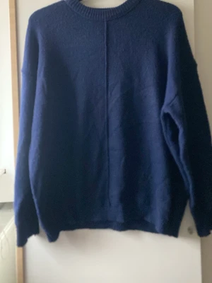 Mörkblå stickad tröja - En enkel och stilren mörkblå stickad tröja med rund halsringning och lång ärm. Tröjan har en rak passform och en diskret söm mitt fram. Perfekt för kyliga dagar och lätt att matcha med olika outfits.