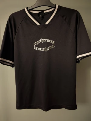 Svart oversized t-shirt H&M XS - Svart oversized t-shirt från H&M med vit text 'togetherness' i gotisk stil på bröstet. V-ringad med breda, ribbade muddar i svart och vitt vid hals och ärmslut. Tillverkad i luftig polyester, perfekt för en chill och sportig vibe.