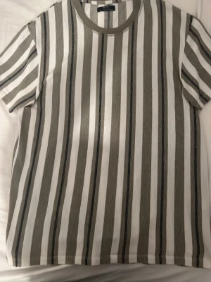 Randig t-shirt från Bershka - Säljer en randig t-shirt från Bershka i storlek L. Skjortan har breda vertikala ränder i vitt, grått och svart samt en rund halsringning med ribbad kant. Materialet känns mjukt och luftigt, perfekt för en avslappnad stil.