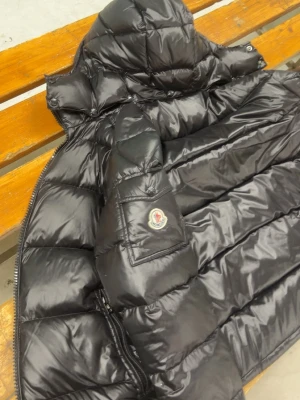 Svart Moncler dunjacka med huva - Säljer en svart Moncler dunjacka med huva och stora quiltade partier. Jackan har dragkedja, ficka med Moncler-logga på ärmen och coolt serietryck på insidan. Perfekt för kalla dagar och riktigt snygg streetstil.. den går att skanna. Storlek S, pris går diskuteras