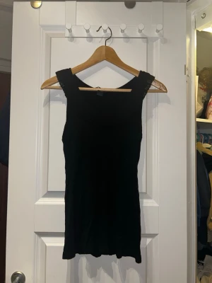 Svart ärmlös topp från H&M - Svart ärmlös topp från H&M med bred, plisserad kant runt halsen. Toppen har en enkel och stilren design, perfekt att matcha med jeans eller kjol. Materialet känns mjukt och lätt mot huden.