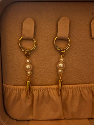 Guldörhängen med pärlor och spets - Säljer ett par snygga örhängen i guldfärg med tre vita pärlor och en spetsig detalj längst ner. Örhängena har klicklås och ger en cool men elegant vibe. Perfekta för att lyfta vilken outfit som helst.