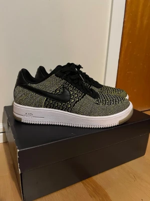 Nike Air Force 1 Ultra Flyknit Low - Säljer ett par Nike Air Force 1 Ultra Flyknit Low i svart och gulgrönt stickat material med vita sulor och svarta läderdetaljer. Skorna har klassisk snörning, svart swoosh och blå detaljer på plösen. Riktigt lätta och snygga sneakers med modern vibe.