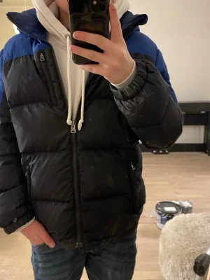 Svart och blå dunjacka Polo Ralph Lauren 152 - Säljer en fet svart och blå dunjacka från Polo Ralph Lauren. Ett fåtal slitningar som man inte lägger märke till. Perfekt för kalla dagar när du vill hålla stilen. Strl 152