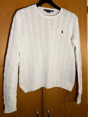 Ralph Lauren Vit kabelstickad tröja  - Kabelstickad vit tröja från Ralph Lauren i 100% bomull. Klassisk rund halsringning, ribbade muddar och ikoniska marinblå loggan broderad på bröstet. Perfekt till jeans eller kjol för en clean och tidlös look.