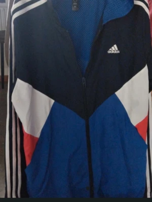 Adidas vindjacka i blått och vitt - Säljer en snygg Adidas vindjacka med färgblock i blått, vitt, svart och röda detaljer. Jackan har dragkedja framtill, meshfoder och klassiska Adidas-ränder längs ärmarna. Perfekt för dig som gillar sportig stil och streetwear.Storlek L ungdom (164)