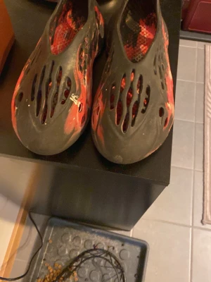 Svarta och röda foam sandaler - Unika sandaler i svart och röd mix med coola utskurna hål över hela skon. Tillverkade i syntetmaterial som gör dem superlätta och bekväma. Perfekta för chill dagar eller när du vill sticka ut med din stil.