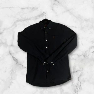 Mörkblå skjorta från Morris - Säljer en stilren mörkblå skjorta från Morris med klassisk krage och vita knappar. Skjortan har lång ärm och liten broderad logga på bröstet. Perfekt för dig som gillar en clean och tidlös look.