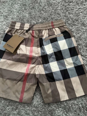 Burberry rutiga shorts med snörning - Snygga shorts från Burberry med klassiskt rutigt mönster i beige, svart, vitt och rött. De har elastisk midja med svart snörning och sidofickor. Perfekta för sommaren och ger en lyxig vibe till din outfit.