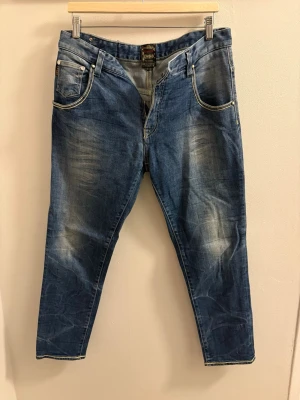 Blå jeans från Rear med kontrastsömmar - Säljer ett par blå jeans från Rear med snygga slitningar och kontrastsömmar. Jeansen har fem fickor, dragkedja och unika detaljer på bakfickorna samt en färgglad patch bak i midjan. Perfekt för dig som gillar en avslappnad men cool stil.