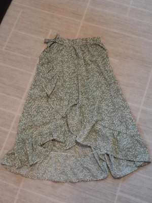 Grön blommig omlottkjol  - Säljer en grön långkjol från SHEIN med vitt blommönster. Kjolen har omlottdesign, volangdetaljer och knyt i midjan. Perfekt för dig som gillar en luftig och somrig stil. Materialet känns lätt och skönt mot huden. !!endast swish +frakt!!