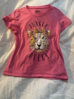 Rosa Jungle Queen t-shirt Juicy Couture - Snygg rosa t-shirt från Juicy Couture med tryck av en leopard med blommor och texten 'Jungle Queen' på bröstet. Klassisk rund halsringning och korta ärmar. Perfekt för dig som vill sticka ut med coolt motiv och färg.