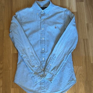 Ralph Lauren skjorta - Säljer denna snygga blå slim fit skjorta från Ralph Lauren i mycket bra skick. Skjortan har långa ärmar och knappar framtill. Den är tillverkad i ett mjukt bomullsmaterial och har en broderad logga på bröstet. Perfekt för en stilren look!