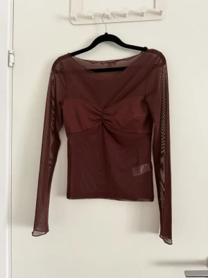 Brun mesh topp - Säljer en brun mesh topp med långa ärmar och rund halsringning. Toppen har en snygg rynkdetalj framtill som ger en cool twist. Materialet är genomskinligt och stretchigt, vilket gör den perfekt att styla lager på lager. Passformen är normal och toppen är lätt croppad. Aldrig använd.