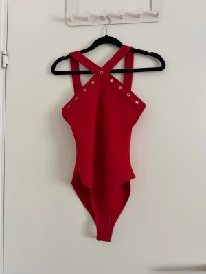 Röd body med silver detalj och korsade band - Säljer en röd body med breda axelband som korsas i ryggen och coola silverfärgade nitar längs överdelen. Västen har en tajt passform och är tillverkad i ett stretchigt syntetmaterial. Perfekt för dig som vill sticka ut med en edgy look. Använd endast för att testas. Storlek S men skulle säga att den passar xs med.